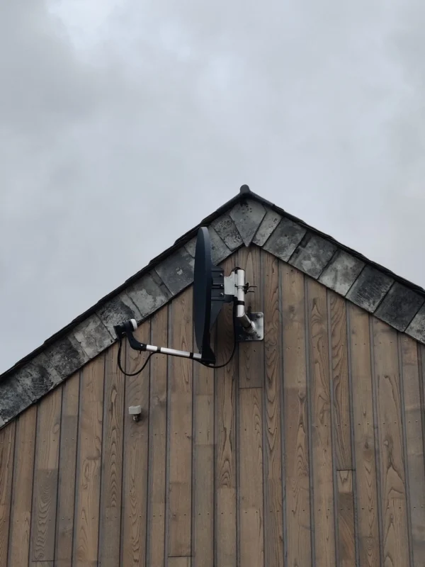 A satellite dish securely mounted on the side of a wooden building with a grey sky in the background.